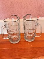 Both clear pressed glass pitchers standing side-by-side on wood surface