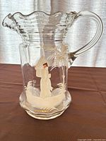 Full view of clear glass pitcher showing ruffled rim, handle, and painted woman