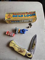 Group shot of all items: folding knife (open), bead loom box, LOVE block, blue hand figurine, metal chain