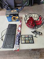 Group shot of all items on table: keyboard, disc repair kit box, packaged flag, hanging basket with liner, fishing reel, stamps