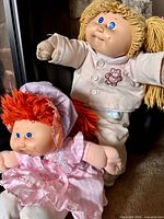 Two Cabbage Patch Kids dolls, blond doll standing behind red-haired doll in pink dress