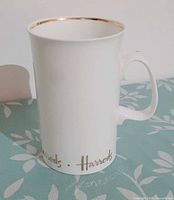 Front view of white Harrods mug showing gold script logo and gold rim