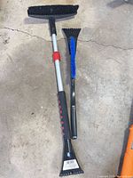 Two car snow brushes on concrete floor: one extendable aluminum brush with scraper, one compact brush with scraper