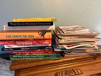 Stack of ten cookbooks and adjacent stack of cooking magazines on wooden surface