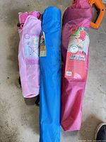 Three folded camp chairs in carry sleeves laid on floor; colours burgundy, blue, pink; label visible on burgundy sleeve
