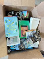 Open box showing assorted books, lantern, blood pressure monitor, tin, media cases and cords