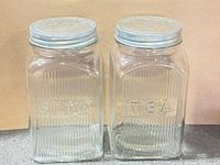 Front view of both ribbed glass canisters labeled SUGAR and TEA with metal lids