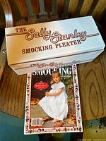 Closed cardboard box labeled The Sally Stanley Smocking Pleater with magazine on table