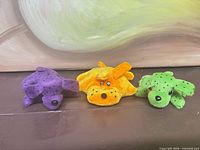 Front view of three plush dogs in purple, orange and green