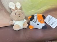 calico cat keychain plush with tag next to tan rabbit plush