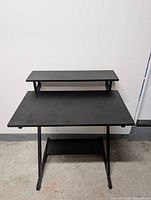 Front view of black computer desk showing main surface, monitor shelf, lower shelf, metal frame