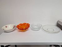 Four serving pieces arranged left to right: Johnson Bros ironstone bowl, orange ruffled glass bowl, clear pressed glass bowl, clear beaded rim platter