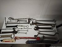 Full layout of assorted wrenches, adjustable wrenches, punch, drill bit, strap and utility knife on tabletop