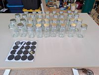 30 square glass spice jars arranged in rows with silver lids; sheet of black circular labels in foreground