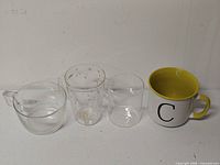 All four mugs lined up: clear round mug, printed star/tree glass mug, plain clear cylindrical mug, ceramic C monogram mug