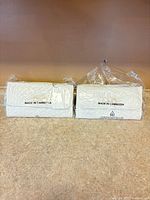 Two identical white faux leather clutches in plastic packaging on countertop