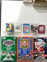 Three Upper Deck factory set boxes (1990 Baseball green box, 1992 Baseball blue/orange box, 1991 NFL Football red box) with several small acrylic card cases containing stickers and promo sets on white tabletop.