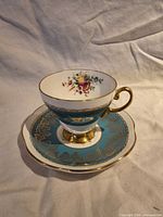 Teacup on saucer showing exterior turquoise band, gold detailing, interior floral basket