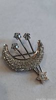 Front view showing crescent moon covered in rhinestones with three star‐tipped rods and dangling star