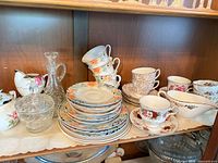 Cabinet shelf showing bone china teacups, saucers, plates, glass decanter, candy dish