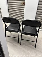 Two black folding chairs open, rear view showing padded backs and seats