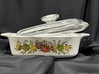 Side profile of casserole dish showing fruit and vegetable motif, lid resting ajar