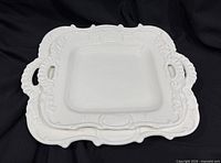 Two stacked square white porcelain trays showing ornate borders and handles