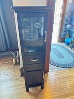 Front view of tall dark brown cabinet showing glass door, drawer, lower door and hardware
