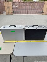 Both closed aluminum cases side by side with measuring tape for scale