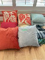 Group view of pillows: coral leaf prints, teal botanical, solid coral velvet, light gray velvet, red knit, plaid throws partially visible