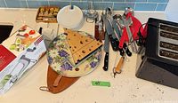 Overview of lot with toaster, boards, platter, knives, utensils, plates, boxed peeler