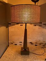 Full view of lit lamp showcasing woven shade, wood column, square base