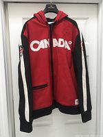 Front view of red Canada jacket showing hood, zipper, pockets and CANADA lettering