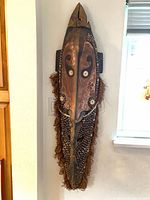Full front view of tall carved wood mask with shells and raffia fringe