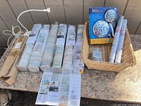 Lot overview showing wallpaper rolls, RainSpa showerhead box, curtain rings pack, window film roll, and boxed shower rod on stone surface