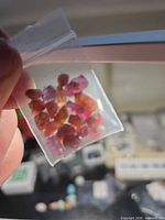 Clear bag holding multiple pink, red and orange rough crystals held between fingers