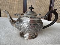 Side view of teapot showing grapevine relief, pedestal base, wooden handle