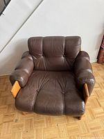 Front view of the brown leather armchair showing seat, backrest, armrests, button tufting and wood supports