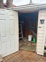 Open shed door showing ladder, long-handled tools, watering can, bucket