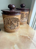 Two stoneware canisters showing relief cabin scenes and maroon lids