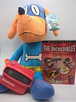 Group shot: Dog Man plush, Incredibles DVD, red View-Master