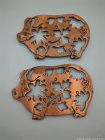 Front view of both pig-shaped trivets showing copper finish and floral cut-outs