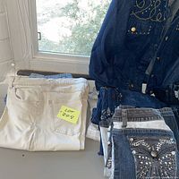 Group shot of folded white jeans, stacked blue jeans, and studded denim jacket