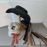 Black leather cowboy hat on stand, beige Rottweiler cap, assorted belts and suede tie accent displayed