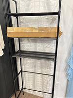 Full view of assembled black metal over-the-toilet shelf with boxed unit resting on shelf