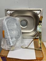 Sink basin, hose assembly in bubble wrap, and wall-mount bracket on top of box