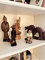 All five figurines on shelf: three wooden figures, ceramic elder, red resin dragon