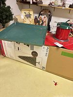 Boxed Ashland 7.5 ft pre-lit tree with green plastic tray and red metal stand on top