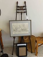 Tall wooden easel holding framed graphite artwork, black frames and woven bag visible