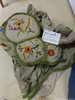 Folded crewel embroidered bed cover with attached tag, partial view of cushions